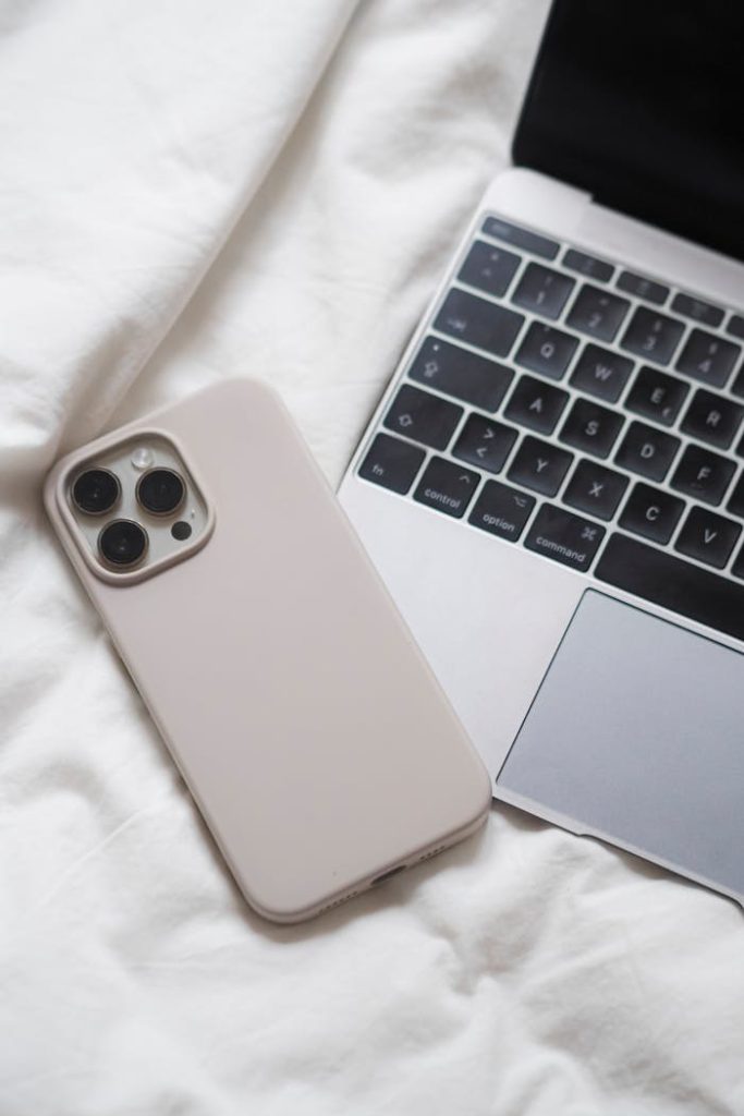 Stylish minimalism: smartphone next to laptop on a white bed, showcasing modern technology essentials.