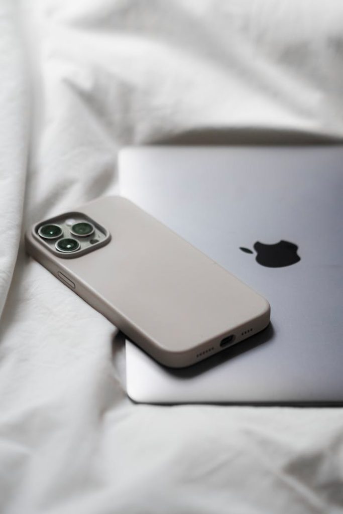 A sleek Apple phone and laptop resting on a white bed sheet, showcasing minimalist design.
